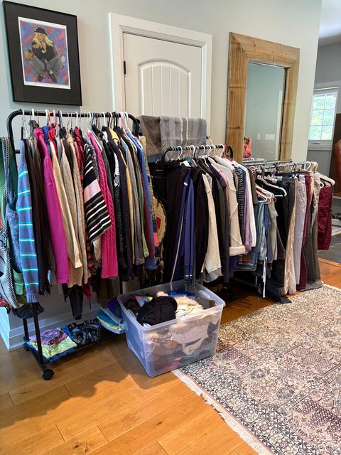 Rails of women's clothing filling a room, with framed art on the wall behind and a bin of smalls on the rug.