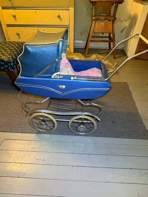 A blue vintage doll's pram with chrome wheels, in front of a yellow painted dresser.