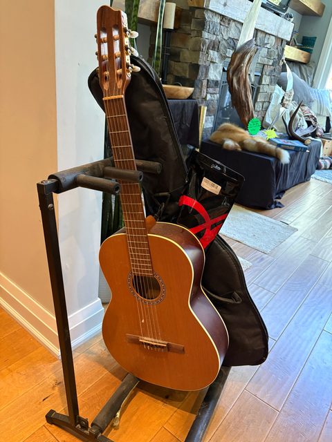 An acoustic guitar on a stand with its soft case leaning behind it.