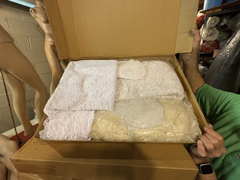 Two hands holding open a cardboard box packed with folded white lace and linens, a dress form standing behind.